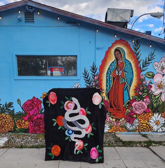 Picture of Laura holding up a snake and rose quilt top in front of a mural that has a blue background with colorful flowers and Our Lady Guadalupe